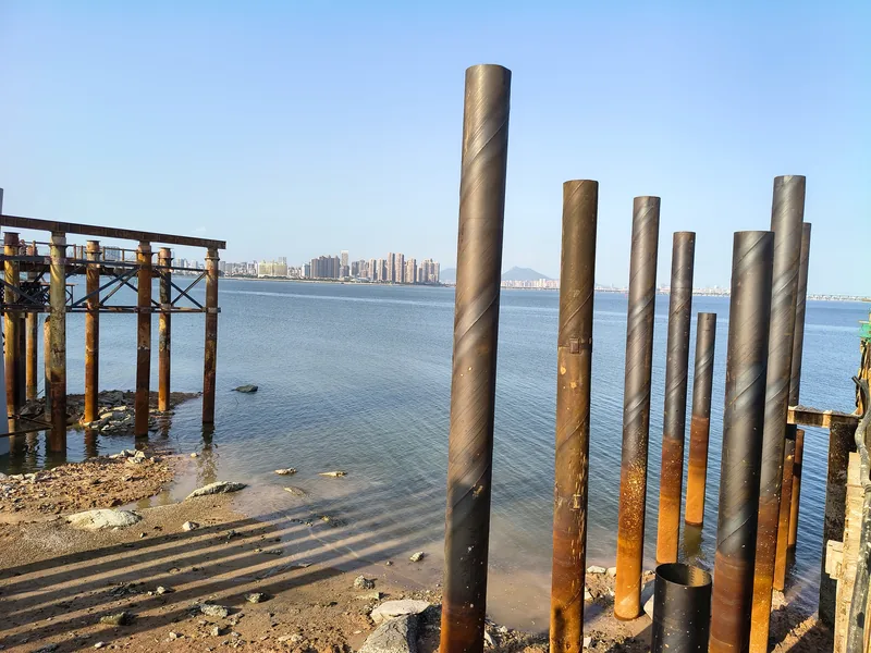 Rows of large-diameter spiral welded steel pipe piles standing upright on a tidal flat, with a braced steel trestle frame on the left and a city skyline across the bay